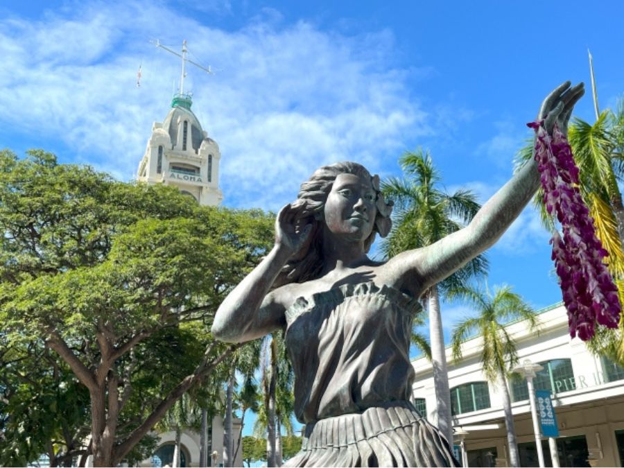 青空に映えるホノルルのアロハタワーと、手前に立つレイをかけたフラダンサーのブロンズ像。