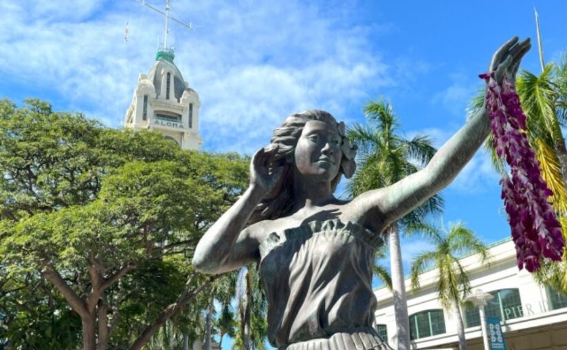 青空に映えるホノルルのアロハタワーと、手前に立つレイをかけたフラダンサーのブロンズ像。