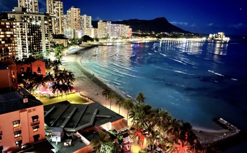 夜のワイキキビーチとダイヤモンドヘッドの全景。立ち並ぶホテルの明かりが海面に反射するホノルルの夜景。